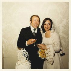 a man and woman standing next to each other in front of a white wall holding wine glasses