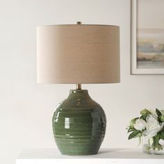 a green vase sitting on top of a white table next to a lamp and flowers