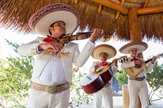 three men in mexican attire playing instruments under a straw hut