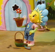 a toy doll holding a potted plant in front of a fake house with flowers