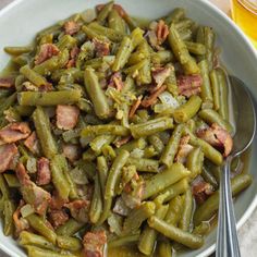 a bowl filled with green beans and bacon next to two silver spoons on a table