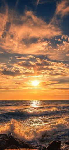 the sun is setting over the ocean with waves crashing in front of it and some rocks