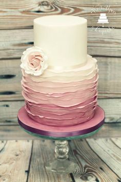 a three tiered cake with pink and white icing on a glass stand against a wooden background