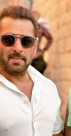 a man wearing sunglasses standing in front of a brick wall and looking at the camera