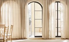 an open window with white curtains and chairs in front of it, next to a dining room table