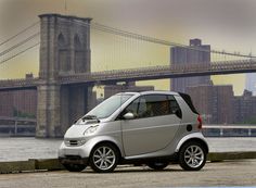 a silver smart car parked in front of a bridge with the city skyline behind it