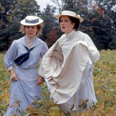 two women in dresses and hats are walking through a field with leaves on the ground