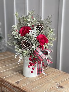 a white vase filled with red roses and greenery on top of a wooden table