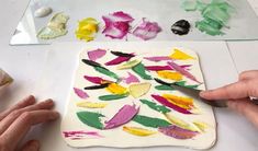 two hands are using scissors to paint on a square piece of paper with colorful flowers