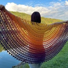 a woman wearing a colorful crochet shawl standing in front of a body of water