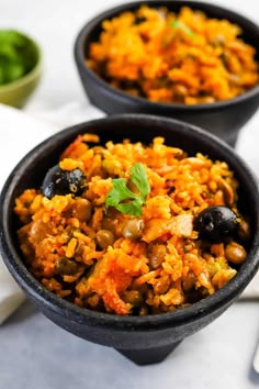 two black bowls filled with rice and olives on top of a white table cloth