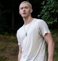 a man in white shirt standing next to some trees and bushes with his hand on his hip