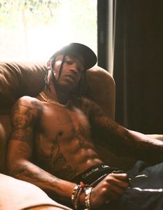 a shirtless man sitting on a couch with his hands in his pockets and wearing a hat