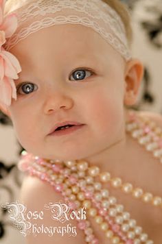 a baby girl wearing pearls and a flower in her hair is looking at the camera