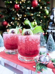 two glasses filled with red liquid and garnished with green olives next to a christmas tree