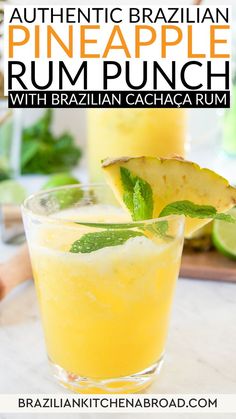 A glass of Brazilian Pineapple Rum Punch topped with a pineapple slice and mint leaves is in the foreground. Behind it, there are fresh limes, a muddler, and a pitcher filled with the same yellow beverage on a white marble surface—perfect for summer.