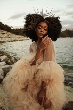 a woman with an afro sitting on rocks near the water wearing a dress and headdress