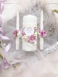 two white candles with pink flowers on them sitting next to purple ribbons and other decorations