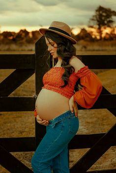 a pregnant woman leaning against a fence with her hands on her hips, wearing jeans and a hat