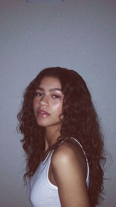 a woman with long curly hair standing in front of a wall and looking at the camera