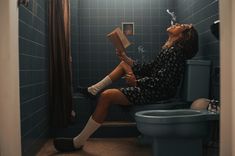 a woman is sitting on the toilet reading a book and drinking from a faucet