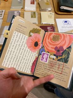 a hand holding an open book on top of a table filled with papers and other items