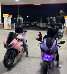 two women sitting on motorcycles in a gas station parking lot at night, one holding the hand of another woman's hand