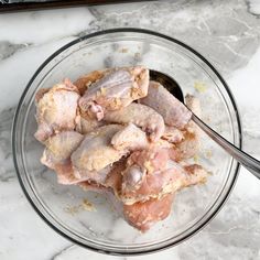 a glass bowl filled with raw chicken sitting on top of a marble counter next to a spoon