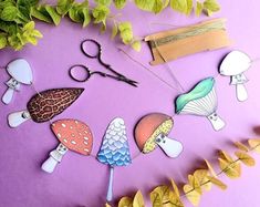 paper cut out of different types of mushrooms on a purple surface next to plants and scissors