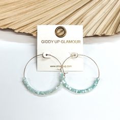 These are thin wire gold hoops with mother of pearl  beads in light blue in the bottom half of the  earrings. These earrings are taken leaning up  against a dried up palm leaf and white background. Giddy Up Glamour, Summer Look, Gold Hoops, Pearl Beads, Summer Looks, Mother Of Pearl, Place Card Holders, Light Blue, Hoop Earrings