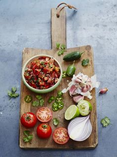 a wooden cutting board topped with tomatoes, onions and green peppers next to a bowl of salsa