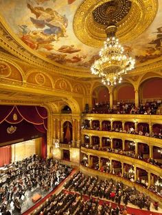 an auditorium filled with people sitting and standing in front of a ceiling painted with gold