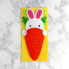 a felt bunny holding a carrot on top of a piece of yellow paper next to a marble surface