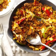 a skillet filled with rice and vegetables on top of a white table next to silverware