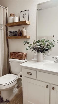 a white toilet sitting next to a sink in a bathroom under a wooden shelf filled with flowers