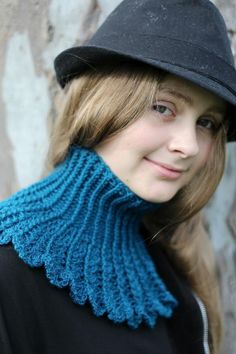 a young woman wearing a black hat and blue knitted neckwarf, standing in front of a stone wall