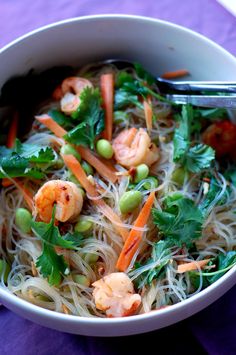 a white bowl filled with noodles, carrots and green vegetables on top of a purple table cloth