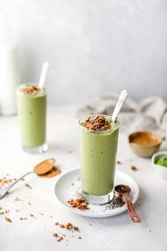 two glasses filled with green smoothie sitting on top of a white plate next to some spoons