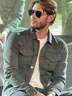 a man with sunglasses sitting down in front of a screen wearing a jacket and jeans