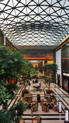 the interior of a shopping mall with tables and chairs, trees, and people walking around