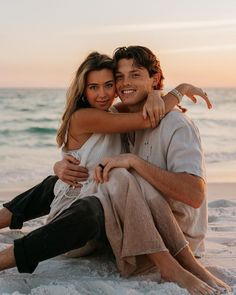 a man and woman sitting on the beach hugging each other with their arms around one another