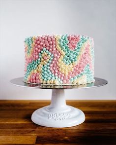 a multicolored cake sitting on top of a white cake plate next to a wooden table