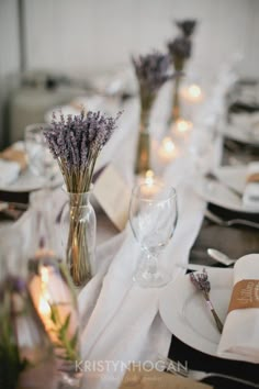 the table is set with white plates, silverware and lavenders in vases