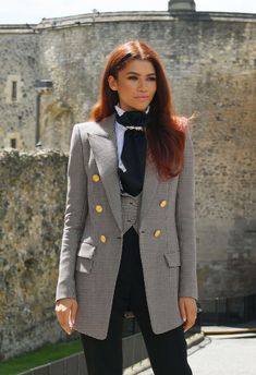 a woman with red hair wearing a suit and black tie standing in front of a castle