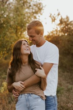 a man and woman are smiling together in the woods at sunset or sunrise, with trees in the background