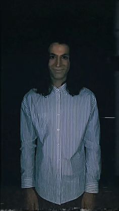 a man standing in the dark with his hands in his pockets and wearing a striped shirt