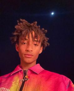 a young man with dreadlocks standing in front of a dark background at night