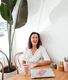 a woman sitting at a wooden table with a laptop and drink in front of her