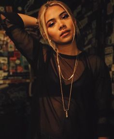 a woman with blonde hair wearing a black shirt and gold necklaces standing in front of a wall