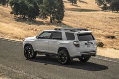 a white toyota 4runner is driving down the road in front of some trees and dry grass